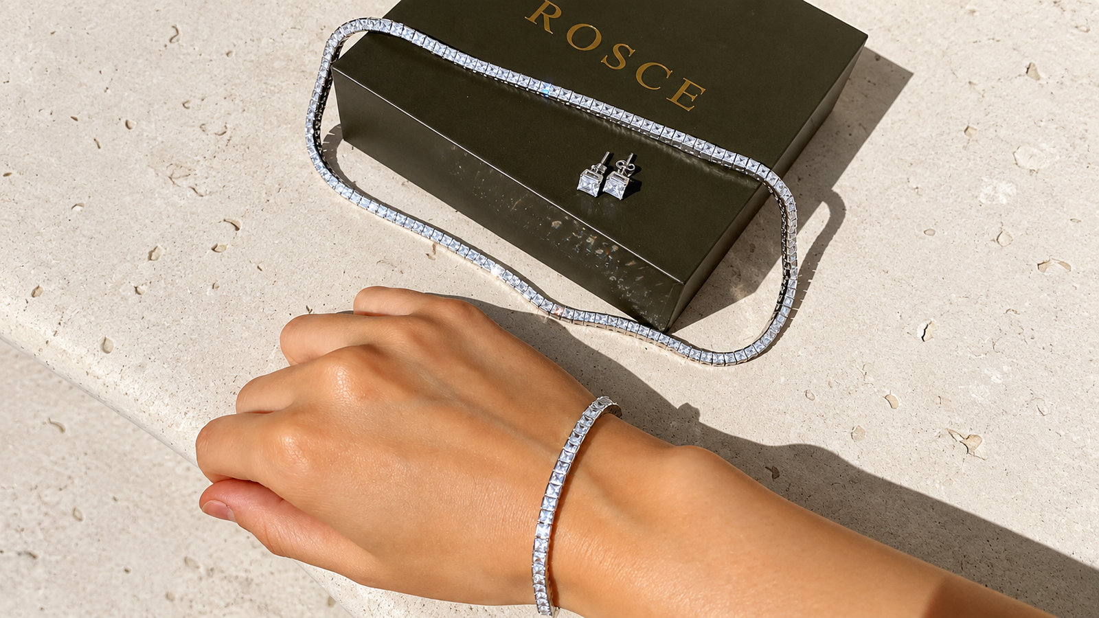 Hand wearing a tennis bracelet beside a ROSCE jewelry box with earrings and a tennis necklace in 18k gold vermeil, showcasing affordable luxury jewelry.