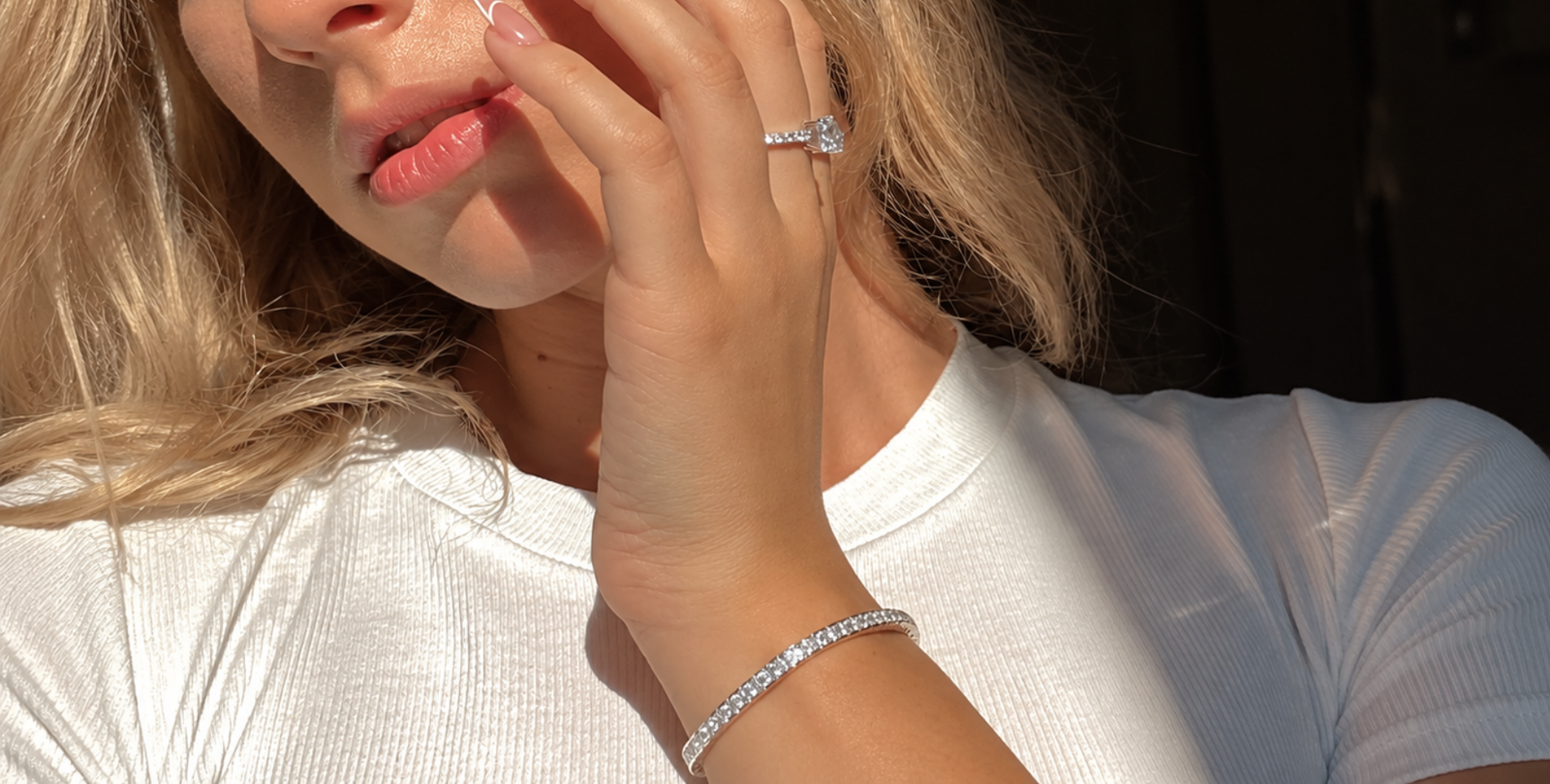 Woman touching her face, showcasing a sparkling tennis bracelet and diamond ring as affordable luxury jewelry.